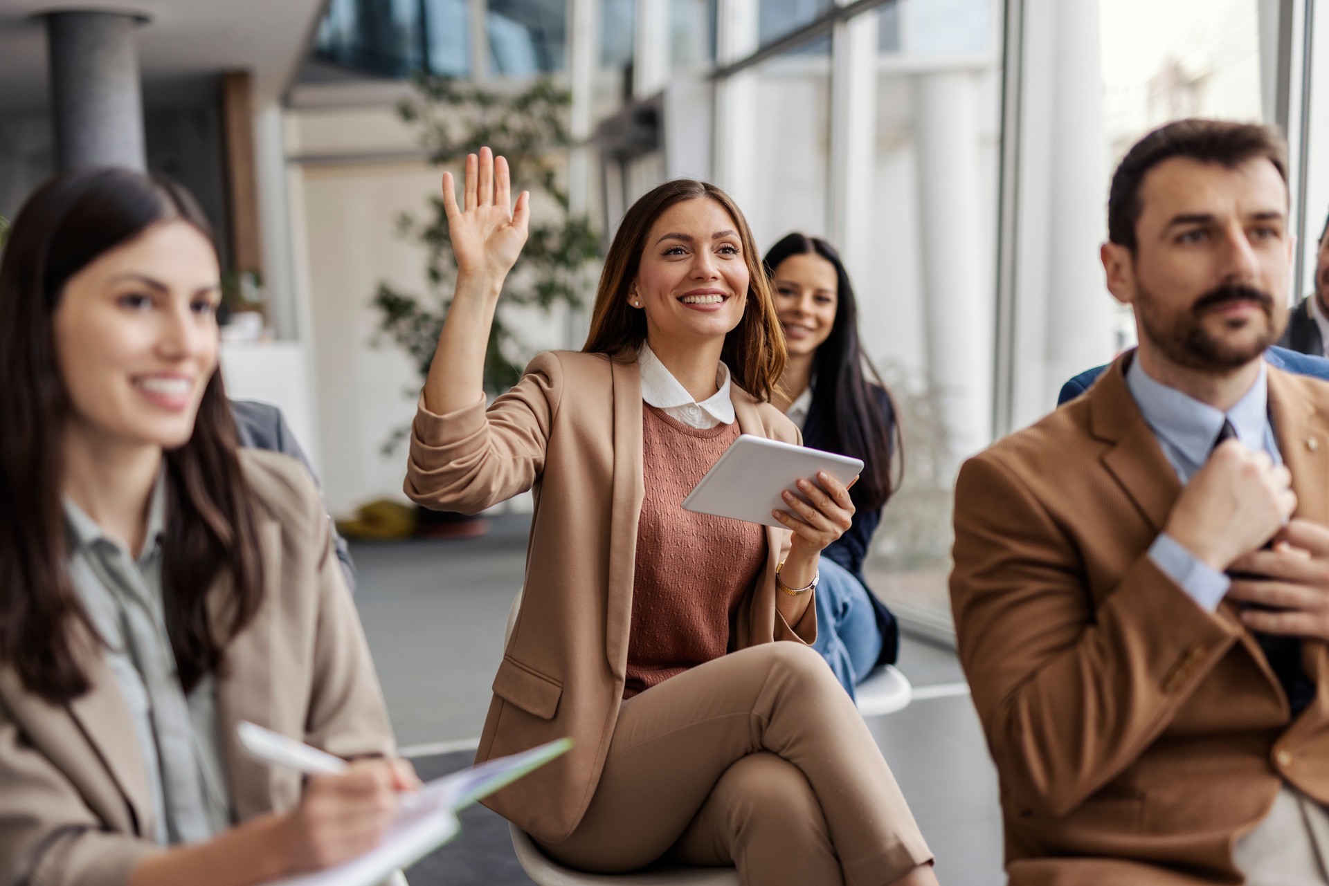 Young professional businesswoman sitting at training room with her colleagues and holding her hand raised.